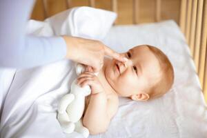 Smiling little kid laying in wooden baby bed under blanket, holding toy, mother finger touching happy kid nose, unrecognizable loving mom playing with awaken cute toddler, closeup photo