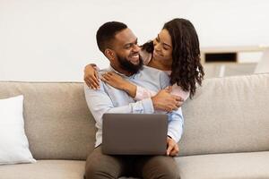 Smiling young black woman embracing man from behind while he using laptop. Loving african american couple spending time together, happy male sitting on the couch working on computer photo