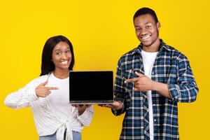 Website ad, online app and self-isolation during COVID-19 lockdown. Cheerful young african american husband and wife hold laptop with blank screen and pointing at it with fingers, on yellow background photo