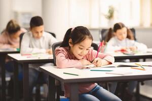 Education And Studying Concept. Portrait of smiling asian small student sitting at desk in classroom, writing in notebook or drawing picture with colorful pencils. Back To School photo