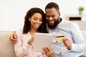 Easy Online Payment Concept. Smiling black couple using digital tablet and showing credit card, choosing and ordering purchases via internet. Woman sitting on the couch, happy man embracing her photo