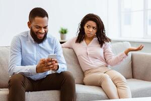 Phubbing. Angry young black woman looking at her boyfriend stuck in smartphone, unhappy about neglect and lack of communication. Jealous lady and gadget addicted man sitting on couch at home photo