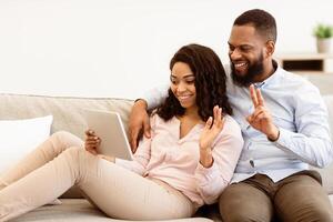 Virtual Communication. Happy black couple having online conference with friends or family using digital tablet, waving hand to web camera, sitting on the sofa in bedroom at home photo