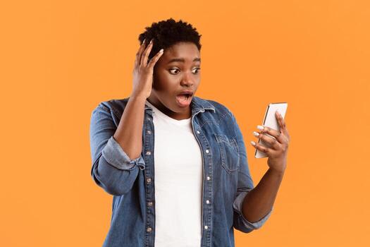 Bad News. Shocked Black Woman Using Phone Touching Head Reading Negative Message Texting Online Standing Over Yellow Studio Background. Mobile Application And Cellphone Problem Concept photo