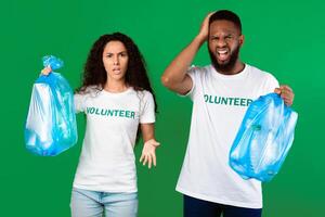 Stop Using Plastic. Two Displeased Multicultural Volunteers Shouting Holding Blue Garbage Bags Full Of Litter Standing On Green Studio Background. Sort Your Waste And Don't Litter Concept photo