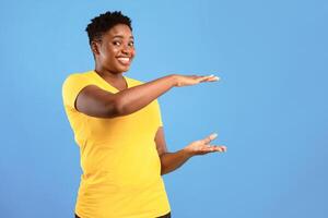 Cheerful Overweight African American Woman Holding Invisible Object Advertising Your Product Or Showing Size Standing Posing Over Blue Studio Background, Smiling To Camera. Copy Space photo