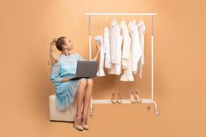 Stylist Concept. Vertical full length portrait of smiling woman sitting on pouffe with laptop computer near rack with clothes and shoes, choosing what to wear, isolated over pastel background photo
