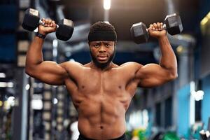 Motivated african american man bodybuilder working on biceps, weight exercises concept. Muscular black guy doing workout with dumbbells at gym, lifting weight up with both hands photo