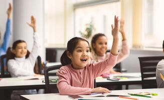 Education, Elementary School, Learning and People Concept. Diverse group of smart junior school kids sitting at desks in classroom and raising hands for an answer, studying with pleasure photo