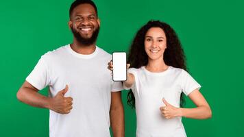 We Like This App. Diverse Couple Showing Smartphone Empty Screen Gesturing Thumbs-Up Approving Mobile Application Standing Over Green Studio Background. Panorama, Mockup photo