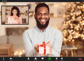 Online Party During Quarantine. Smiling african american bearded guy making online call, showing present gift box to his surprised happy girlfriend at videochat. Screen view with conference app photo