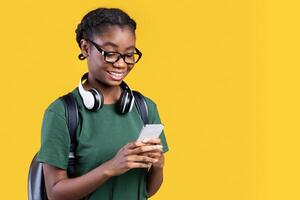 Happy African Female Student Using Smartphone Networking And Texting Online Standing Over Yellow Background. Young Woman Using Mobile Application On Phone. Studio Shot photo