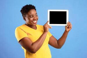 Smiling African American Woman Showing Digital Tablet Blank Screen Advertising Application Or Website Standing Over Blue Studio Background. Female Holding Computer With Empty Monitor. Mockup photo