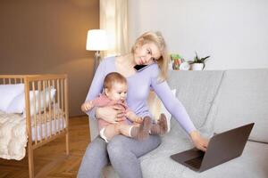 Stressed working young mother with little baby on her lap having business conversation on mobile phone, looking at laptop screen, home interior, copy space. Working mothers concept photo