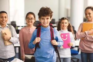 Return Back To School. Smiling small boy wearing backpack posing in classroom, diverse group of happy international classmates standing in line in the blurred background, studying and learning photo