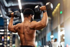 Back view of african american shirtless man bodybuilder with muscular body exercising at gym, lifting up dumbbells, black man making work out for his biceps and back, copy space photo