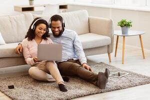 Virtual Communication. Happy black couple in headphones having online conference with friends or family using computer, waving hand to web camera, sitting on the floor carpet in bedroom at home photo