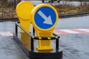 A bright yellow directional sign with a blue circular background and white arrow points left in a rainy urban setting. The sign is placed on a pedestrian crossing area made of black paving stones photo