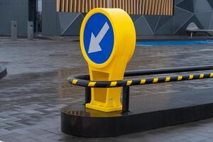 A bold yellow directional sign with a blue circular background indicating leftward movement stands on a sleek pavement in an urban setting. The surroundings showcase contemporary architecture photo