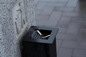 A black waste container filled with discarded cigarette butts stands on a wet pavement under gray skies in a bustling city photo