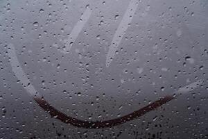Raindrops softly cascade down a foggy window, creating a serene atmosphere. A playful smiley face drawn through the mist brings a sense of warmth and joy to an otherwise dreary day photo