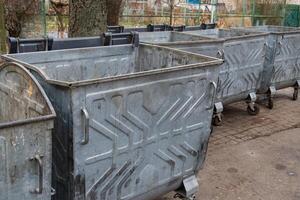 Several large, metallic garbage containers stand side by side on a paved surface, surrounded by trees photo