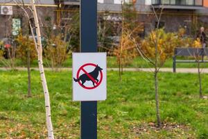 A bright warning sign displays a crossed-out dog symbol, indicating that dogs are not allowed to relieve themselves in this green public area. The backdrop features autumn foliage and urban elements photo