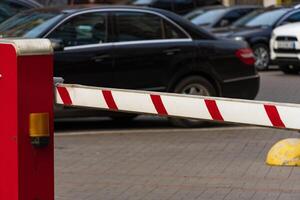 A bright red barrier stands at the entrance of a busy parking lot, preventing entry while vehicles wait in line. The scene reflects measures for safety and security in public spaces photo