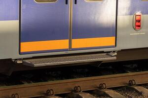 Close-up of a modern electric train door positioned at a railway station. The vibrant colors contrast with the rail tracks and surrounding environment during sunset, creating a striking visual photo