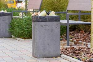 A concrete garbage container is overflowing with trash bags and recyclables in a well-maintained urban park. Nearby benches and greenery enhance the outdoor atmosphere, highlighting waste management photo