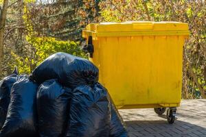 A yellow garbage container stands next to multiple black trash bags piled on a paved area photo