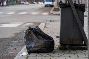Two black trash bags rest beside a metallic garbage container along a city sidewalk, emphasizing the ongoing issues with urban waste disposal and environmental conservation efforts photo