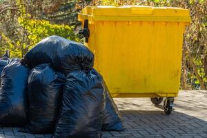 A yellow garbage container sits beside several black trash bags on a paved surface outdoors. The scene captures the importance of proper waste disposal and recycling efforts in the community photo