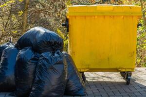 Black garbage bags are piled beside a bright yellow waste bin in a park. The surrounding trees suggest it's daytime, emphasizing the need for environmental cleanliness and proper waste disposal photo