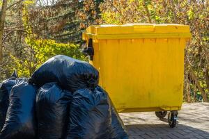 Several black garbage bags are piled next to a yellow waste container in a residential outdoor setting. The greenery around adds to the environmental context photo