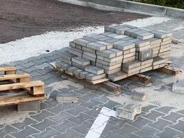 Stacked paving bricks rest on wooden pallets at a construction site where a sidewalk is being laid. Various materials are scattered around, indicating ongoing work photo
