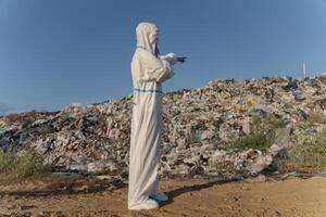 A person in protective gear stands at a trash dump, pointing towards a large pile of garbage. The surrounding area reveals the extent of environmental pollution from human activities photo