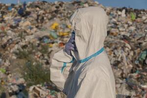 A worker in protective clothing stands amid a large landfill filled with mountains of garbage, visibly engaged in assessing the pollution and its effects on the ecosystem photo