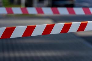 Caution tape with bold red and white stripes stretches across a pathway, clearly marking a restricted area in a busy city setting. The sun casts shadows on the pavement photo