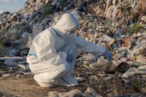 A researcher in protective clothing is crouching at a landfill, analyzing waste and pollutants in a puddle photo