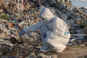 An environmental worker in protective gear carefully inspects polluted water at a landfill. The area is filled with plastic debris, reflecting concerns about waste and sanitation photo