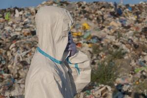 A worker in protective gear stands at a landfill, surveying a large area filled with trash and plastic waste under a clear sky. This highlights issues of pollution and ecology photo
