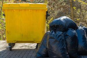 Bright yellow trash bin stands prominently alongside a mound of black garbage bags in a peaceful outdoor location. Sunlight filters through surrounding trees, creating a vivid scene photo