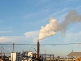 A factory stands tall with smoke rising from its chimney as the sun sets, casting a warm glow over the industrial scene. The sky remains predominantly blue, highlighting the structure photo