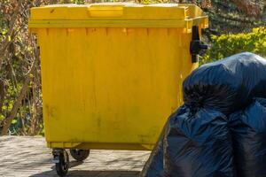 The yellow garbage bin is parked beside several black trash bags, surrounded by vibrant greenery, hinting at an afternoon of waste collection in the neighborhood photo