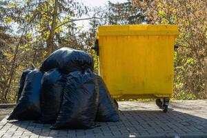 A bright yellow dumpster is placed next to a large pile of black garbage bags, illuminated by warm autumn sunlight filtering through green and brown leaves photo