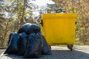 A bright yellow garbage can contrasts against dark plastic bags, set against a backdrop of trees showcasing the colors of fall on a sunny day photo