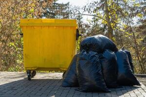 A striking yellow garbage can rests next to a heap of black garbage bags, set against a backdrop of colorful autumn leaves and clear skies, reflecting a quiet urban moment photo