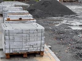 Stacks of gray construction materials rest on wooden pallets beside a muddy area, surrounded by gravel and soil piles under a gloomy sky, ready for the ongoing project photo