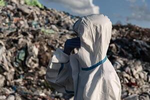 A worker clad in a protective suit inspects a sprawling landfill, surrounded by piles of waste under a partly cloudy sky. The significance of environmental safety is evident photo